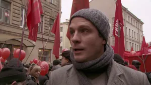 Foto van een man, je ziet zijn gezicht, in een betoging/demonstratie met veel mensen die rode vlaggen en rode ballonnen dragen.