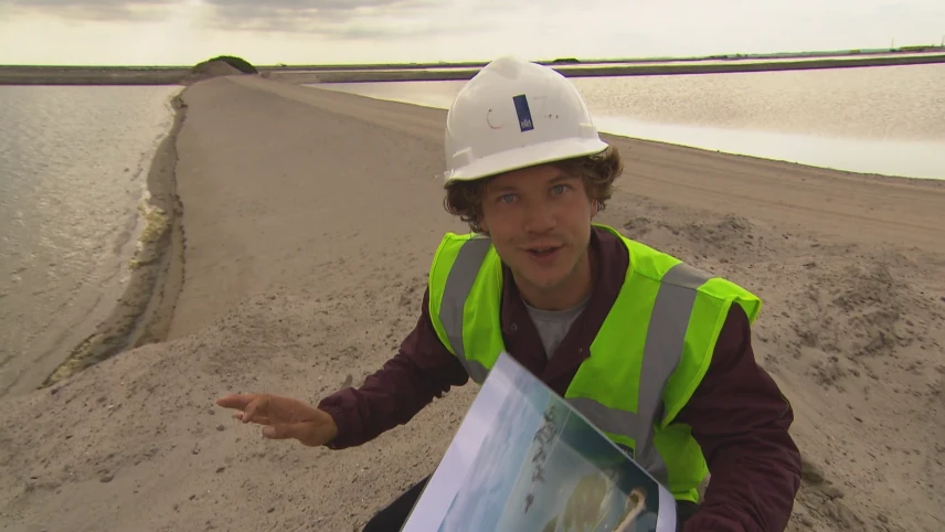 Foto van presentator in werkkleding met een kaart in de handen die staat bij een lange pas aangelegde zanddijk in het water.
