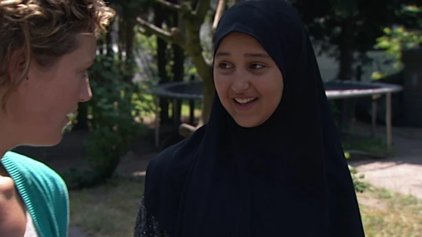 Foto van gesluierde vrouw in gesprek met presentator, buiten in een tuin met mooi weer.