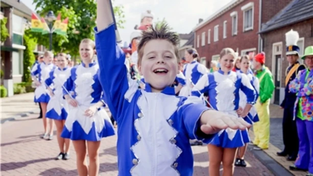 Foto van majorettes, kinderen, blauw/wit gekleed, die in optocht door een straat lopen, voorop loopt vrolijk jongetje, de aanvoerder.