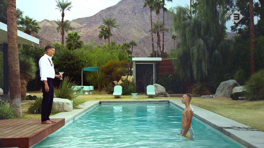 Foto van naakte man die staat in zwembad in luxe tuin met palmen, naast het zwembad staat net geklede man met glas drank in zijn hand.