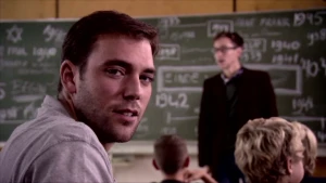 Foto van man, je ziet zijn hoofd, in een klas, je ziet nog paar kinderen, de leraar die staat voor het schoolbord.