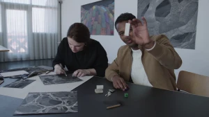 Man en vrouw die aan een grote tafel zitten waarop tekeningen liggen, de man houdt een gum vast en laat deze zo zien.