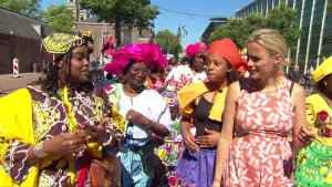 Foto van aantal vrouwen en presentator op straat in een stad, de vrouwen zijn vrolijk gekleed, vieren Keti Koti.