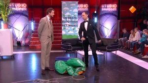 Foto van de studio van Snugger waarop de presentator staat met een andere man, op de grond ligt een groene plastic zak met een touw eraan.