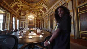 Foto van presentator die staat in grote zaal, de Treveszaal, met grote tafel erin met veel stoelen eraan.