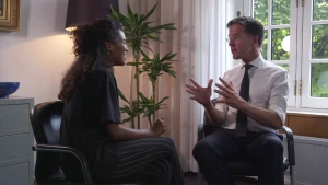 Foto van premier Mark Rutten die staat in kamer, van het Torentje, en praat tegen een vrouw die op een stoel zit.