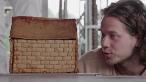 Foto van het hoofd van presentator die kijkt naar een modelletje van een stevige muur van een huis.