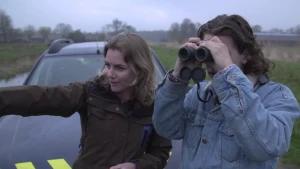 Foto van man en vrouw, de man kijkt door een verrekijker, bij een auto in de natuur.