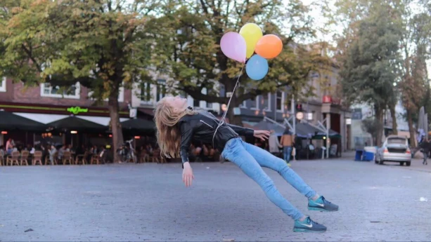 Foto van meisje op een stadsplein dat achterover heldt, om haar middel zit een touw met aantal ballonnen eraan.