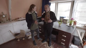 Foto van presentator en oude man die in een moderne huiskamer staan met een bureau.