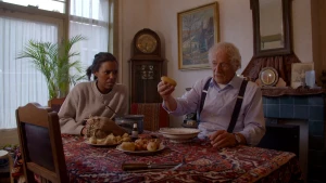 Foto van presentator en oude man die aan tafel met eten erop zitten in huiskamer.
