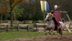 Foto van man verkleed als Middeleeuwse ridder op een rennend paard met schild en lans op renbaan.