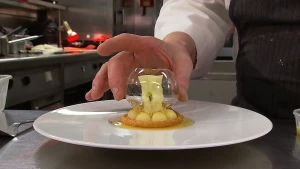 Foto van een bord met een mooi bijzonder opgemaakt toetje erop en hand van kok die er iets mee doet.