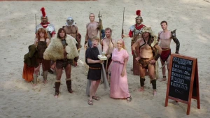 Foto van aantal mensen en kinderen verkleed als Romeinen met wapens zoals een lans, staan op het zand van het Colosseum.