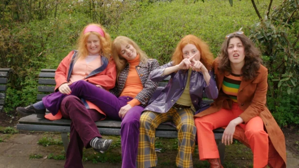 Een foto van vier vrouwen in kleding met oranje, paarse, roze, gele en groene kleuren. Ze zitten buiten op een houten bankje met erachter groene planten of struiken. Eén van de vrouwen legt haar wijsvingers tegen elkaar, zodat ze een letter "X" vormen. 