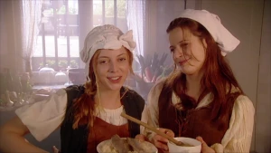 Een foto van twee vrouwen in een keuken met een wit kapje op hun hoofd. De ene vrouw heeft een wit bord vast met zoute haringen erop die op zuurkool liggen. De andere vrouw heeft een wit kommetje vast, met een houten lepel erin, waar zuurkoolsoep inzit. 