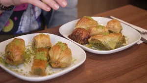 Een foto van een bruine tafel met Janouk's hand erboven en erop stukken baklava op twee witte borden. De baklava bestaat uit bladerdeeg met groene stukjes op de buitenkant. Er zitten pistachenootjes in met niet-zichtbare eieren, boter en suikersiroop. 