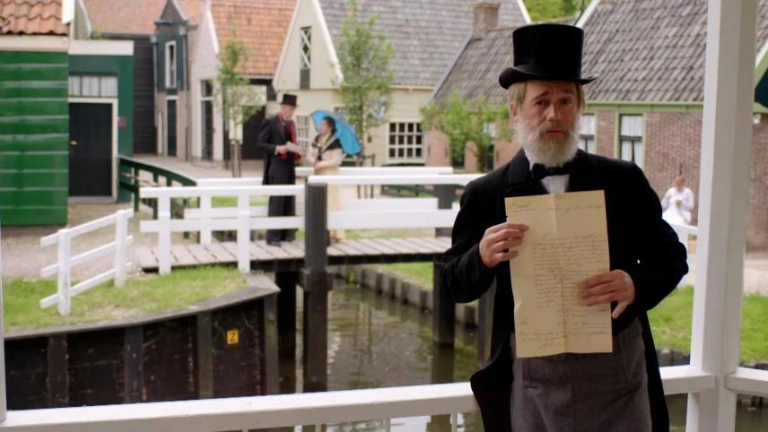 Een foto van een man verkleed als Sam van Houten, in een zwart pak, met een zwarte hoed en een zwarte strik om. De man heeft een geelachtig document vast en staat op een soort brug met witte leuningen. Erachter liggen water, een witte brug en huizen.