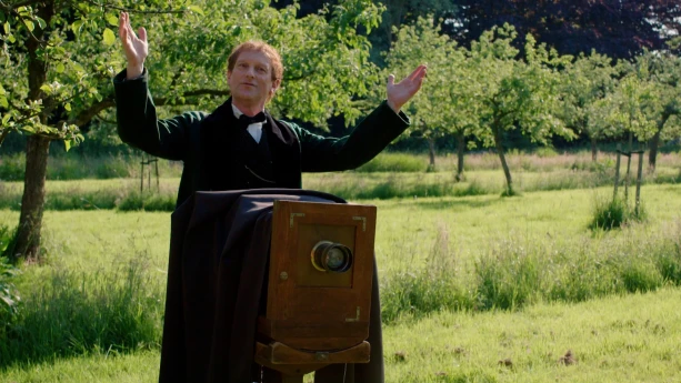 Een foto van een man verkleed als fotograaf Joseph Niépce die op een groen grasveld staat achter een heel oud fototoestel. Het fototoestel is een soort rechthoekige, houten kast met een ronde, zwarte lens erin. Achter de kast hangt een donker doek. 