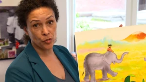 Een foto van schrijver en tekenaar van kinderboeken, Mylo Freeman. Ze heeft een tekening vast van prinses Arabella die een gouden kroontje en een oranje jurk draagt en op het hoofd van een grijze olifant zit. De olifant loopt op een gelige grasgrond.