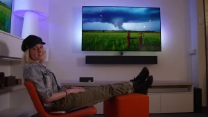 Een foto van presentator Janouk die in de camera kijkt terwijl ze achterover in een stoel zit. Op de achtergrond is er een televisie te zien die aanstaat.