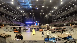 Een foto van een ruimte, een concertzaal, die opgebouwd wordt door mensen.  Een deel van een podium is opgezet in het midden, er ligt een stapel planken en metalen palen, er staan kisten en apparaten met schuifjes, knopjes en beeldschermen op een tafel.