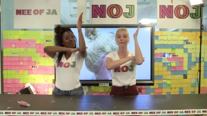 Een foto van Sosha en Janouk achter een tafel, met hun linkerarm omhoog en hun rechterhand op hun bovenarm, direct achter de omhoogwijzende linkerarm. Op een beeldscherm staat een foto van de zijkant van Janouk's hoofd, haar nek en haar zijn hier te zien.