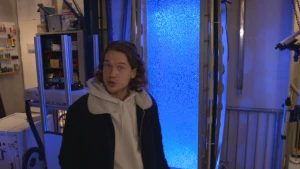 Een foto van Pascal die naast een rechthoekige, hoge bak staat met metalen buizen aan beide zichtbare zijkanten. In de bak zijn allemaal bubbels te zien in het water. De bak wordt verlicht door blauw licht.