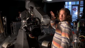 Een foto van Pascal die bij een grote camera staat. De camera zit met gekleurde kabels aan een grote, metalen draaiarm en de grote, ronde zwarte lens wijst iets naar beneden in de richting van de camera die gebruikt is om deze foto te nemen. 