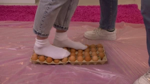 Een foto van de benen van Janouk en Sosha. Janouk loopt met witte sokken over vier kartonnen dozen met bruine eieren heen. Eén voet staat op één rij eieren, de andere voet staat voor de helft op twee rijen. Eronder ligt plastic over het paarse vloerkleed.