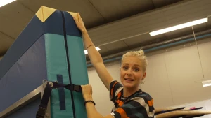 Een foto van Janouk die een metalen rek met een dikke mat die gebruikt wordt voor gymnastiek erop vastheeft in een grote ruimte. De mat zit door een zwarte band vast op het rek. De mat is groen van kleur met geelachtige hoeken.