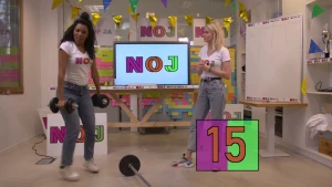 Een foto van Sosha die twee gewichten in haar handen heeft en naast haar lichaam houdt en een klein beetje omhoog springt. Janouk staat naast een wit bord en kijkt naar Sosha. Op de grond ligt een gewicht met een lange, metalen buis als handvat.  