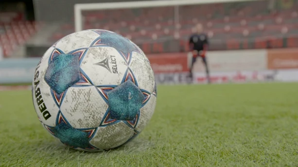 Een foto van een voetbal met witte en blauwe vlakken op een groen grasveld. Op de achtergrond is onscherp een stadion zonder publiek te zien en daarvoor een doel met een keeper in het midden. De bal ligt klaar voor het nemen van een strafschop.