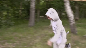 Een foto van Janouk die naar links rent in een bos. Ze draagt een onesie, een pak dat aan elkaar zit. De onesie is een soort grijze hamster of muis met een witte buik met grijze strepen erop en met een hamsterkop als capuchon.