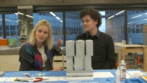 Een foto van Janouk en een man achter een tafel. Op de tafel staat een voorbeeld of maquette van een hoog, rechthoekig, grijsachtig kantoorgebouw of flatgebouw met acht rechthoekige blokken, die voor een deel op elkaar staan en deels in de lucht hangen.