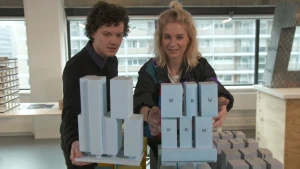 Een foto van een man en Janouk die achter een rechthoekig ding staan met hoge, rechthoekige, blokken erop. Het zijn blauwgrijze voorbeelden van een gebouw, maquettes. Bij één maquette staat een zwarte letter op ieder blok, een "W" een "K"  of een "H".