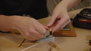 Een foto van de handen van iemand achter een lichtbruine tafel, waarop stukjes door midden gespleten bamboehout liggen en dubbelgevouwen bamboehout op een soort stokje. De persoon heeft een stukje opgevouwen bamboehout vast, dat op een soort stokje zit.