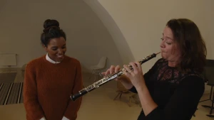 Een foto van een vrouw die speelt op een hobo, een zwart, houten blaasinstrument. Het is een lange buis met zilverkleurige knoppen en buisjes erop en er steekt een staafje uit, het mondstuk waar de vrouw op blaast. Sosha staat er met een blije lach naast.