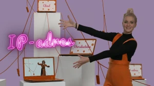 Een foto van Janouk die met beide handen wijst naar het roze woord "IP-adres" voor de foto. Er staan vier hoge, witte rechthoeken met oranje draden eromheen en laptops erop. Op één laptop staat iemand met opgeheven handen en een masker voor in een badjas.