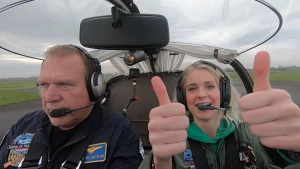 Een foto van Janouk en een man die in een rondom doorzichtig vliegtuigje zitten, dat nog op de landingsbaan staat of net begonnen is met opstijgen. Ze dragen een hoofdtelefoon met microfoon. Janouk houdt twee duimen omhoog en kijkt naar de camera.