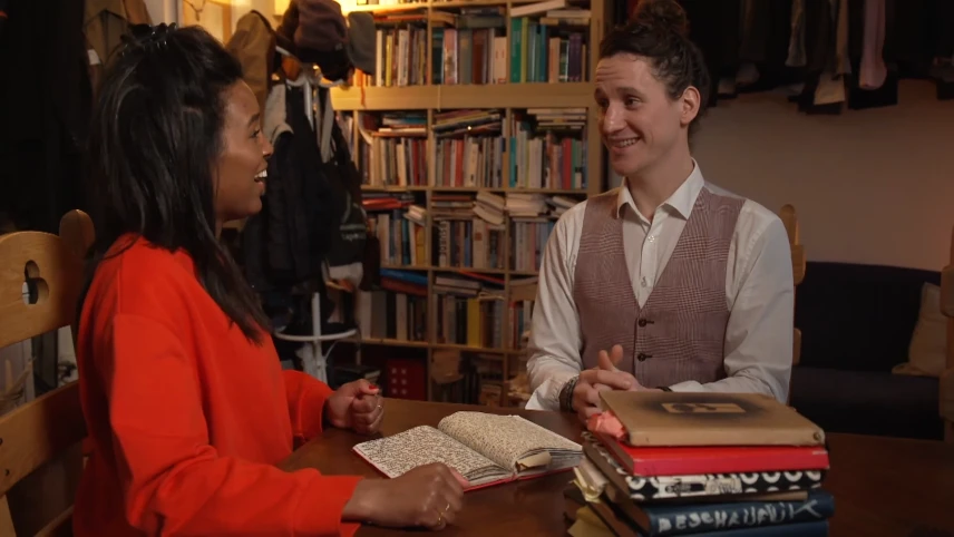 Een foto van Sosha en Jesse die achter een tafel staan. Op de tafel ligt een stapel boeken en ligt een opengeslagen boekje vol met geschreven tekst. Sosha en Jesse lachen naar elkaar. Op de achtergrond staat een boekenkast vol met boeken.