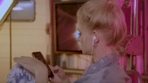 Een foto van Janouk die zit op een soort stoel met opgetrokken knieën. Ze heeft witte oordopjes in, die in de telefoon zitten die ze in haar hand heeft. Haar andere hand hangt boven haar knie.