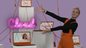 Een foto van Janouk die met beide handen wijst naar het roze woord "Cloud" links. Er staan vier hoge, witte rechthoeken met oranje draden eromheen en daarop laptops met de woorden "cloud", "versleutelen" en "ip-adres" erop en iemand verkleed als een wolk.