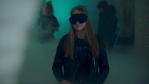 Een foto van een meisje met een zwart oogmasker voor haar ogen. Ze staat in een groenachtig verlichte ruimte met witte wolken op de vloer en links naast haar. Op de achtergrond lopen twee mensen naar de rug van het meisje toe.