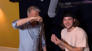 Een foto van Pascal die in een ruimte staat met een andere man. Een grote, grijze microfoon hangt boven ze in de lucht. De man ernaast heeft lange, bruine slierten of linten in zijn hand en maakt er ritselende geluiden mee.