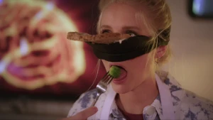 Een foto van Janouk die een zwarte band om haar hoofd heeft die onder haar ogen, om haar neus, zit. Er ligt een bruine boterham op, waar pindakaas bovenop zit. Profs hand heeft een vork vast en stopt daarmee een groen spruitje in de mond van Janouk.