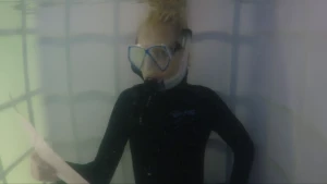 Een foto van Janouk in een soort bak met water. Ze heeft een donkergekleurd pak aan en draagt een blauwe en deels doorzichtige duikbril, waarvan een doorzichtige, plastic slang loopt naar een zwart mondstuk. Ze leest van een wit vel papier in haar hand.