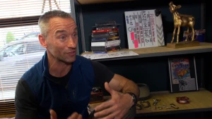 Een foto van een man naast een muur met planken. Op de planken ligt een stapel doosjes van films op DVD, een filmklapper of wit bord dat dichtklapt zodat acteurs weten dat filmen begint met "Floris en de heilige nagel" erop, en de filmprijs gouden kalf.