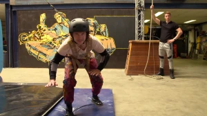 Een foto van Pascal die door zijn knieën gebogen op een blauwe mat staat. Hij draagt een zwarte helm en heeft een tuigje om zijn lichaam, waar een touw aanzit dat omhoog loopt. Rechts staat een man die een touw vastheeft dat naar boven loopt.